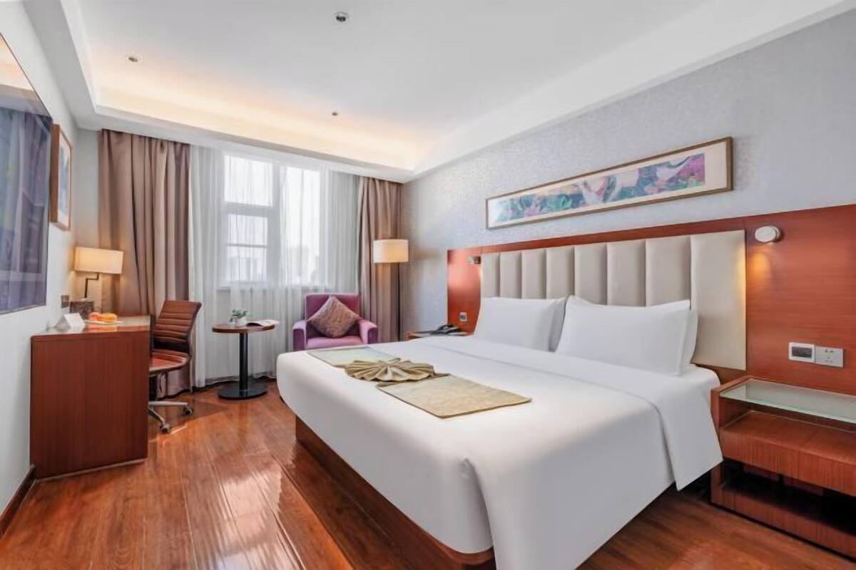 Quarto de hotel com uma cama grande, uma escrivaninha, uma cadeira, uma mesa pequena e uma janela com cortinas.