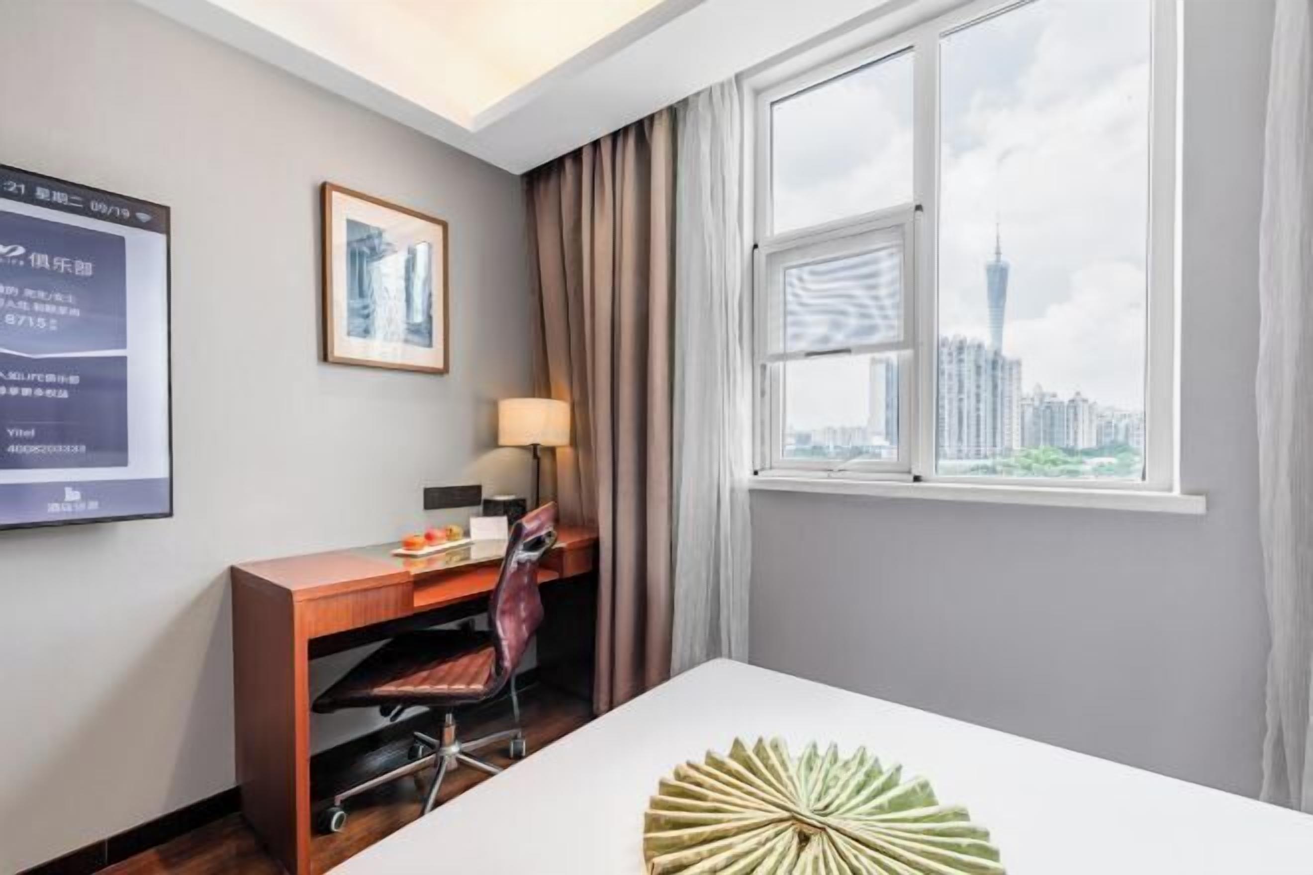Quarto de hotel com escrivaninha, cadeira, luminária e vista para a cidade através de uma grande janela.
