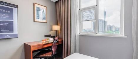 Quarto de hotel com escrivaninha, cadeira, luminária e vista para a cidade através de uma grande janela.