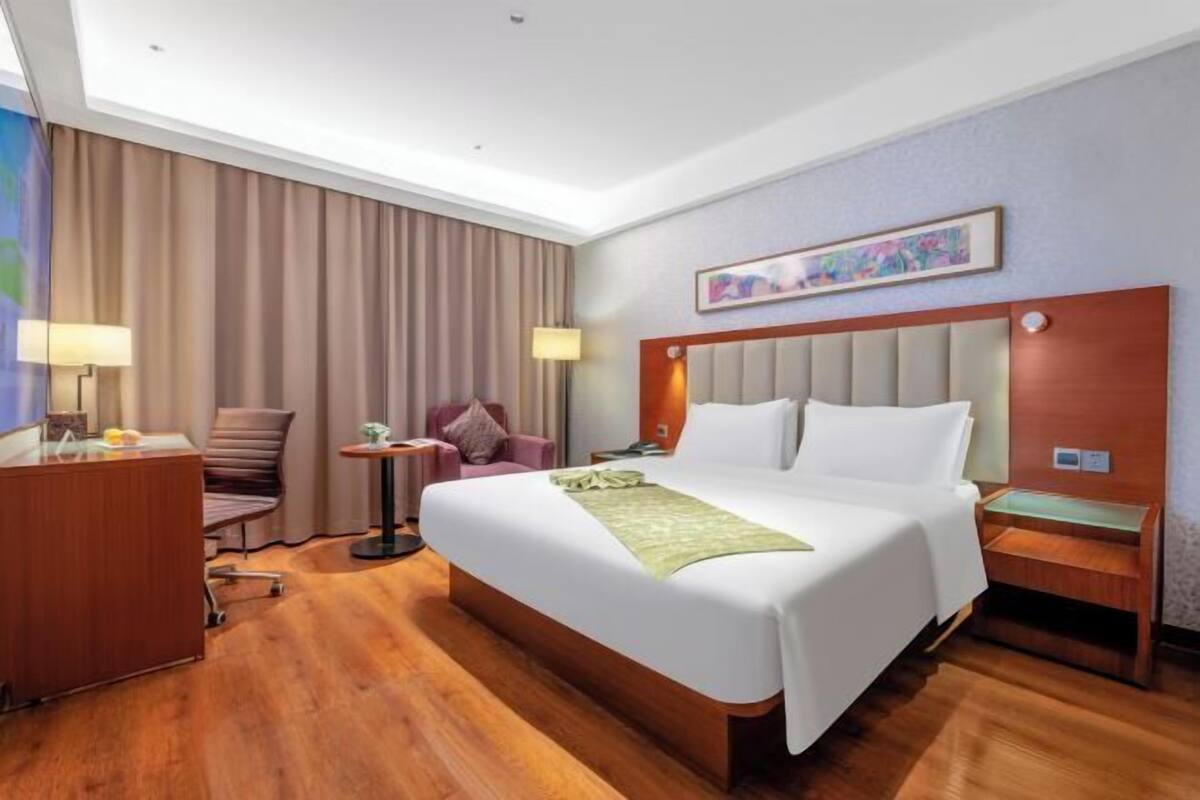 Quarto de hotel com uma cama grande, uma escrivaninha, uma cadeira, uma mesa de cabeceira e um quadro na parede.