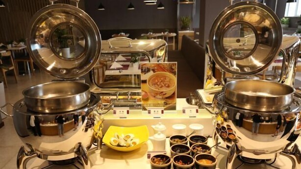 Un'attrezzatura per buffet con diverse postazioni di servizio in acciaio inox, un'esposizione centrale con cibi e un'area salotto sullo sfondo.