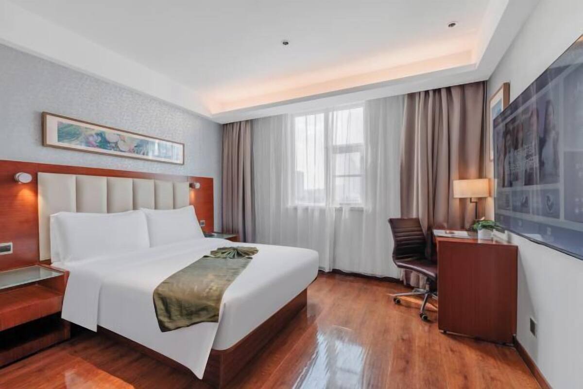 Quarto de hotel com cama, escrivaninha, cadeira, luminária e uma janela grande com cortinas.