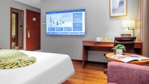 Una camera d'albergo con un letto, una scrivania, una sedia, una televisione a parete e un quadro incorniciato sopra la scrivania.