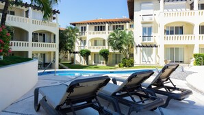 A poolside area with lounge chairs, a swimming pool, and a building with balconies and windows.