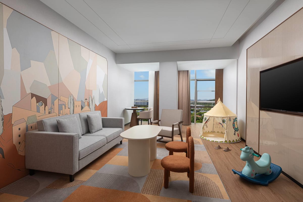 Une chambre d’hôtel moderne, équipée d’un canapé, de fauteuils, d’une petite table, d’un téléviseur et d’une fresque murale représentant un paysage urbain.