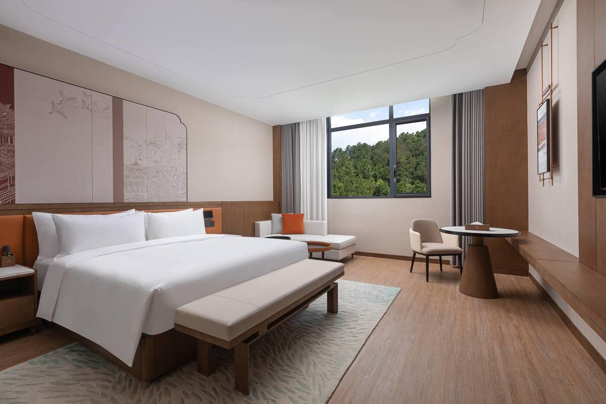 Une chambre d’hôtel moderne dotée d’un grand lit, d’une table de chevet, d’une chaise, d’une petite table ronde et d’une grande fenêtre donnant sur un paysage verdoyant.