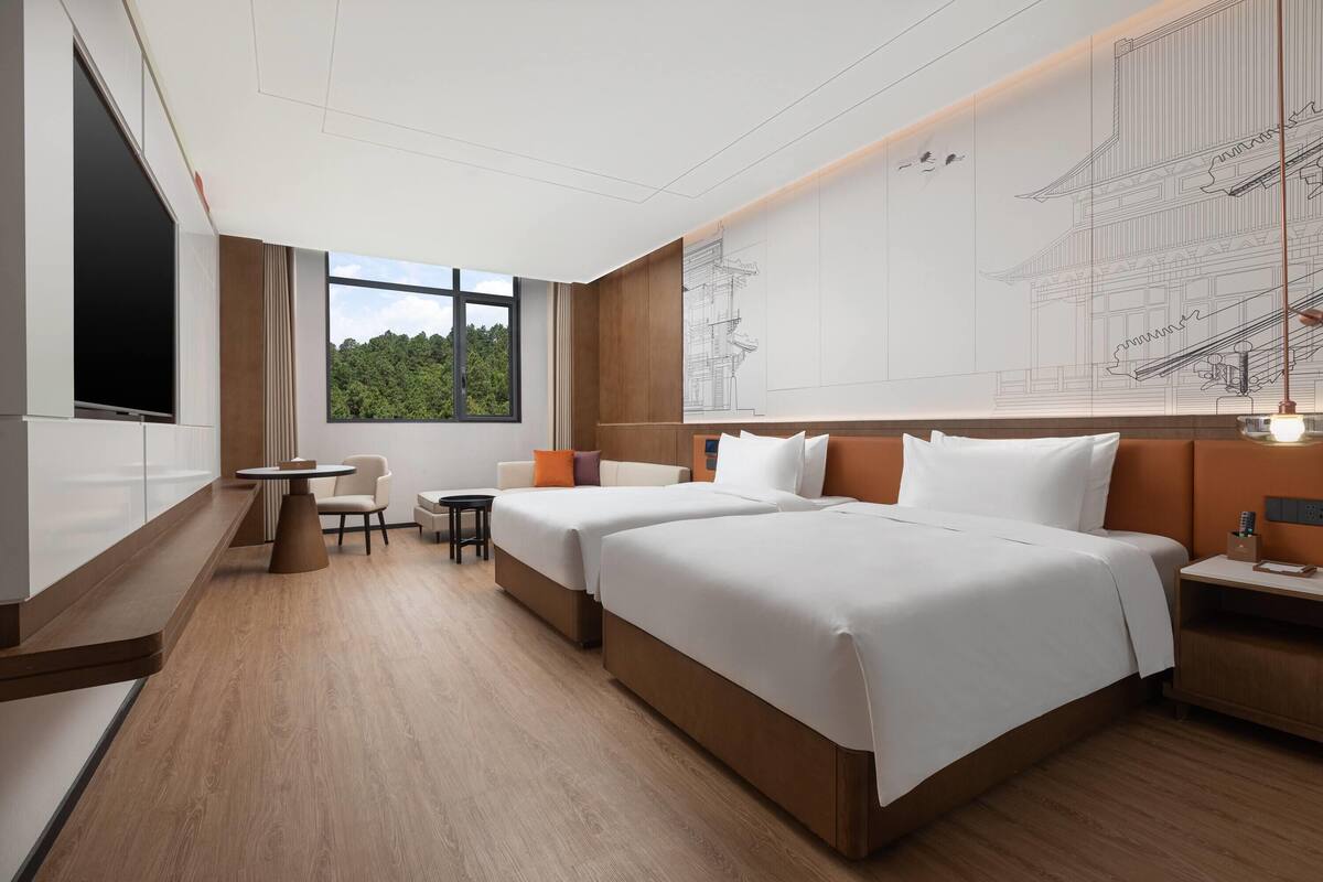 Une chambre d’hôtel avec deux lits, un coin salon avec une table et des chaises, une grande fenêtre donnant sur un espace verdoyant et un téléviseur fixé au mur.