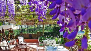An outdoor patio with white metal chairs and tables, surrounded by greenery and purple flowers. - La Corte No.3 (Massa Lubrense)