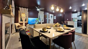 Un comedor moderno con una mesa blanca dispuesta para cuatro personas, un sofá y un televisor al fondo.