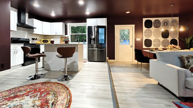 Una cocina moderna con armarios blancos, un refrigerador de acero inoxidable y un microondas. Hay dos taburetes de bar y una mesa de comedor con sillas. Las paredes están pintadas en tonos oscuros, y hay una pared decorativa con diseños circulares.