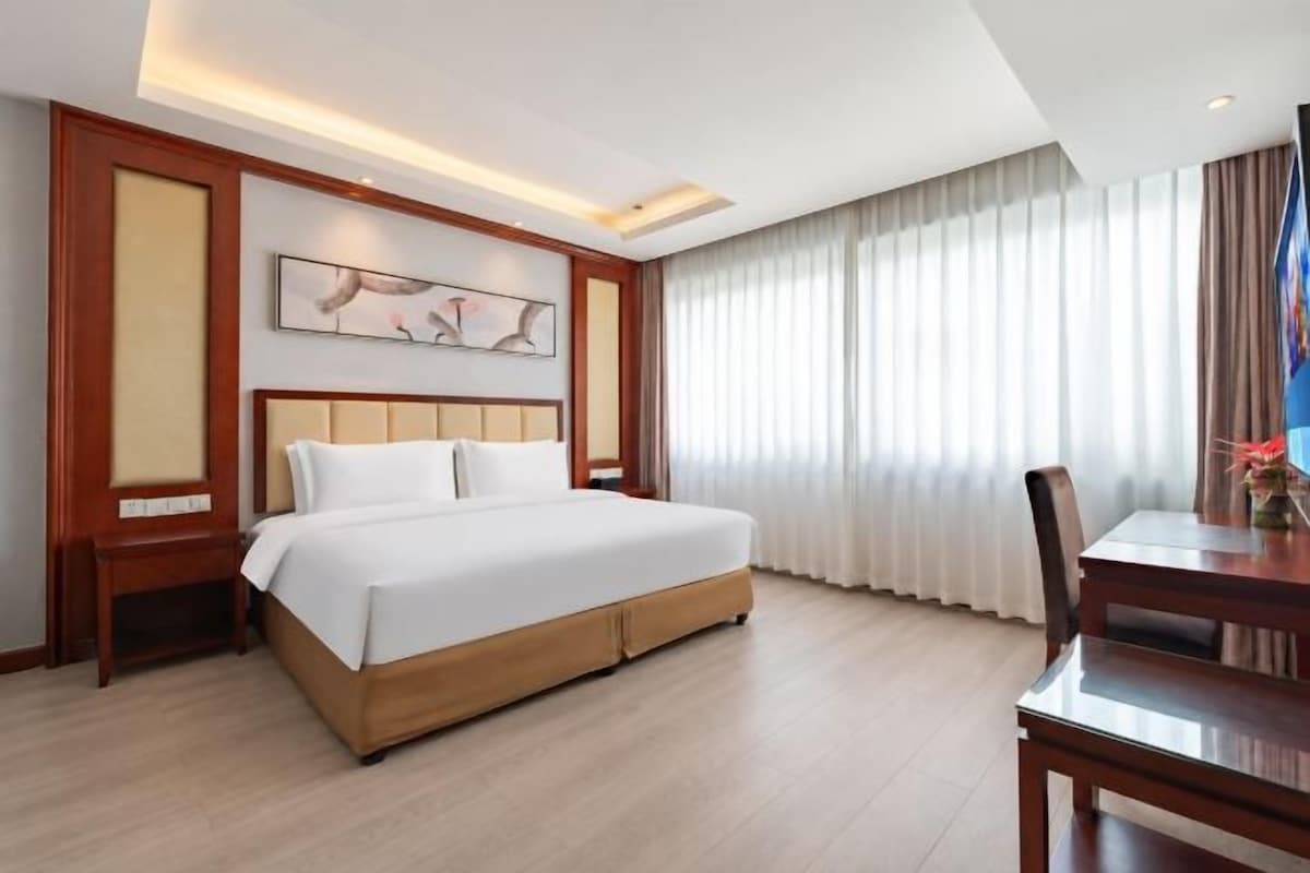 Une chambre d’hôtel avec un grand lit, une tête de lit en bois, une table à manger et une vue sur les rideaux.