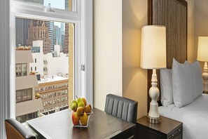 Une chambre d’hôtel avec une table à manger, un panier de fruits, une lampe et un lit avec des oreillers.