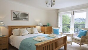 Ein Schlafzimmer mit einem hölzernen Bett, Nachttischen, einem Sessel, einem Balkon mit Aussicht und einer Chandelüster.