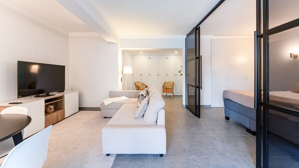 A modern living room with a flat-screen TV, a sofa, a dining table, and a kitchen area with cabinets.