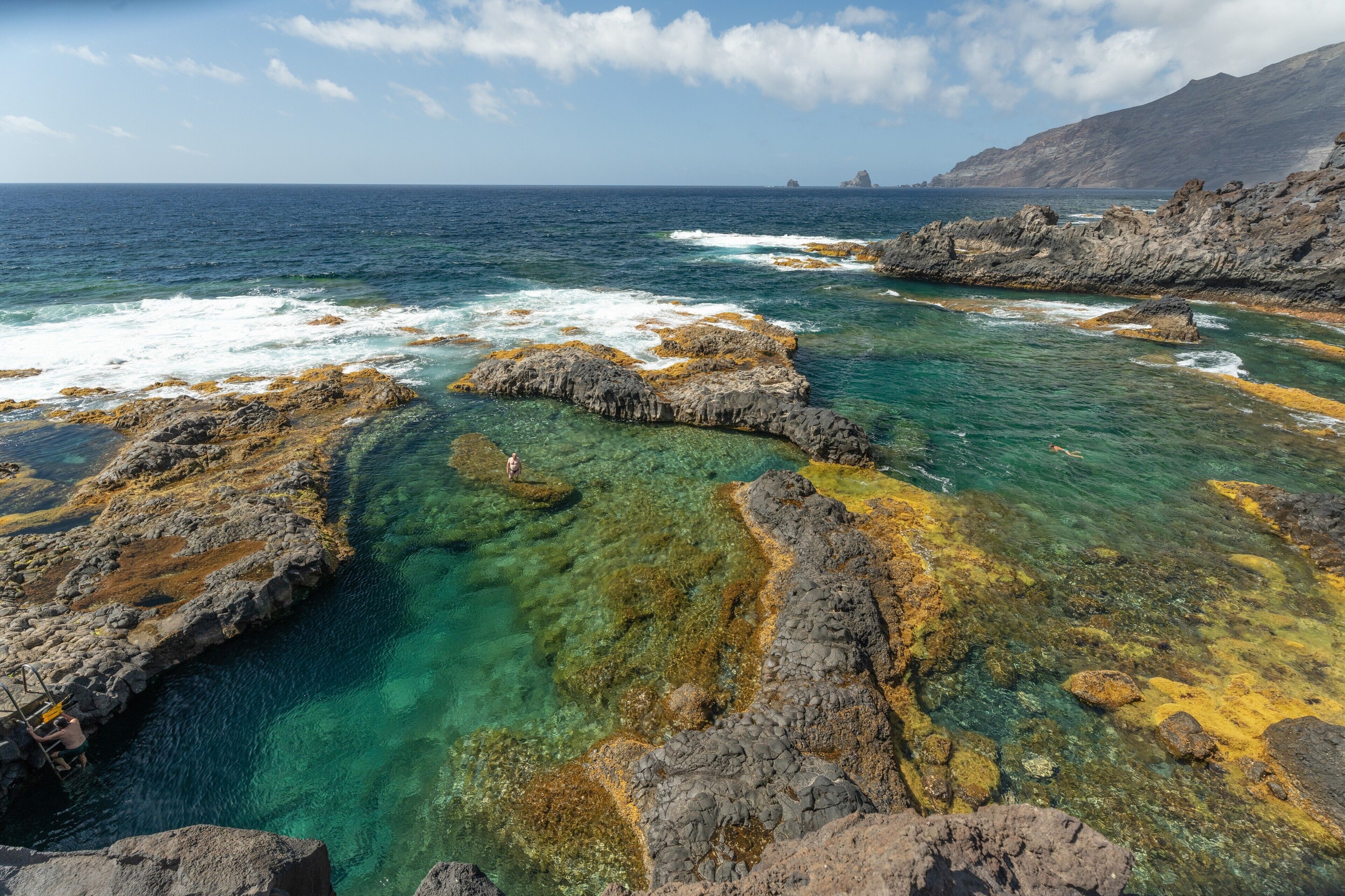 Un paysage côtier caractérisé par des eaux limpides, des formations rocheuses et des montagnes à l’horizon.