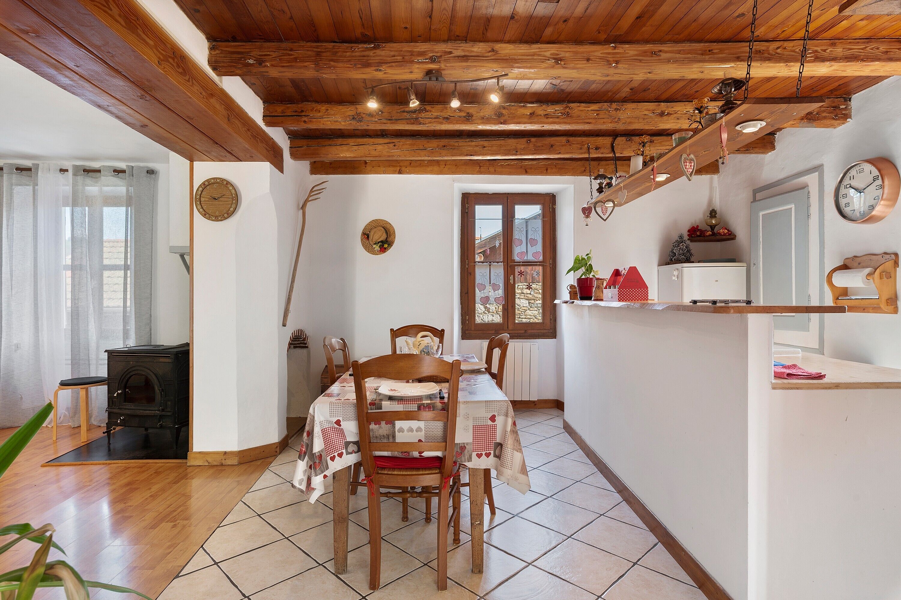 A rustic kitchen with wooden beams, a dining table, chairs, a stove, and a window with curtains.
