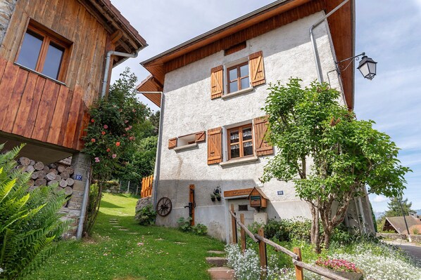 A rustic building with wooden shutters, a stone wall, and a garden with flowers and a wooden fence.