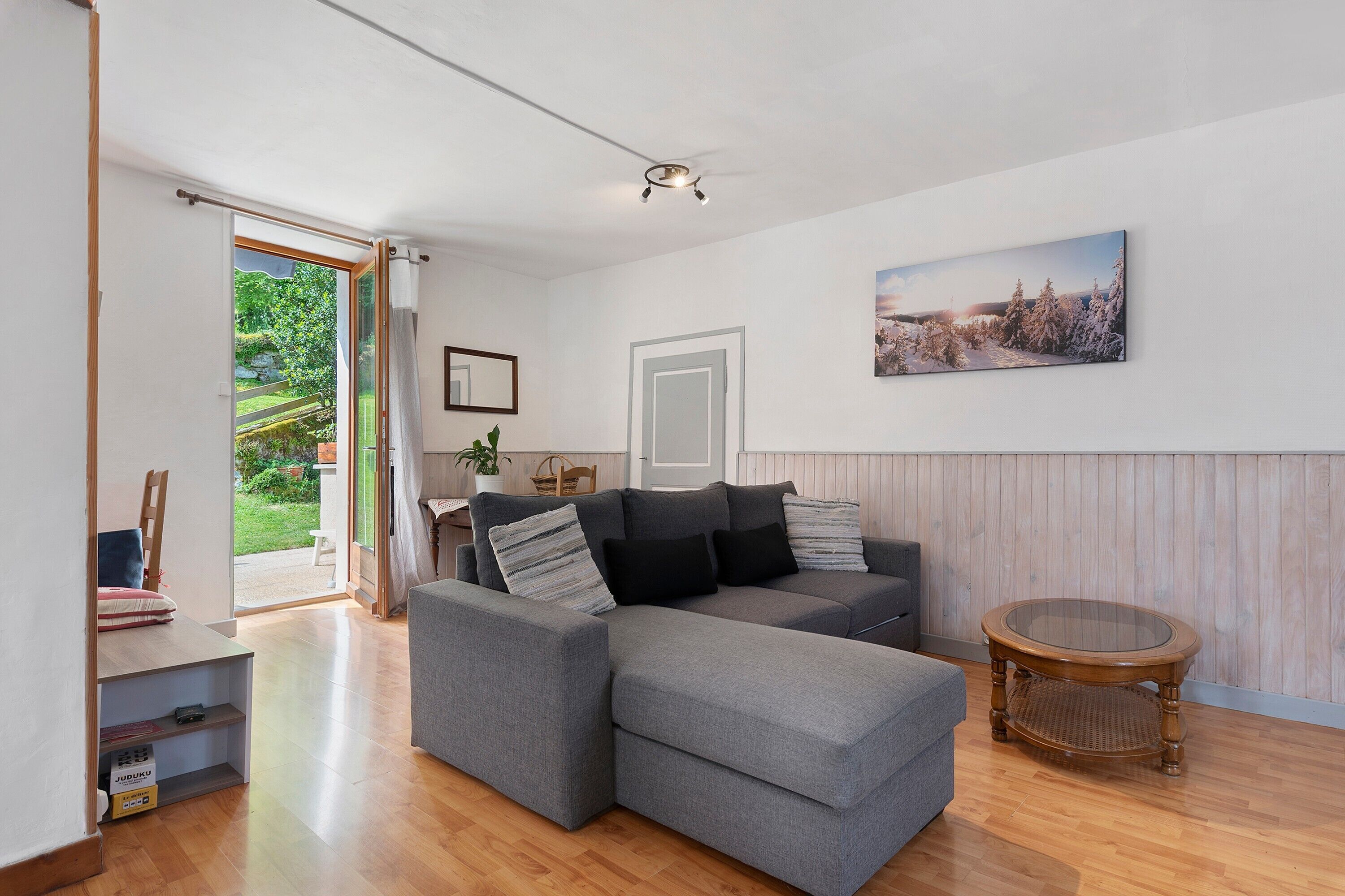 A modern living room with a grey sectional sofa, a round wooden coffee table, and a large window offering a view of greenery.