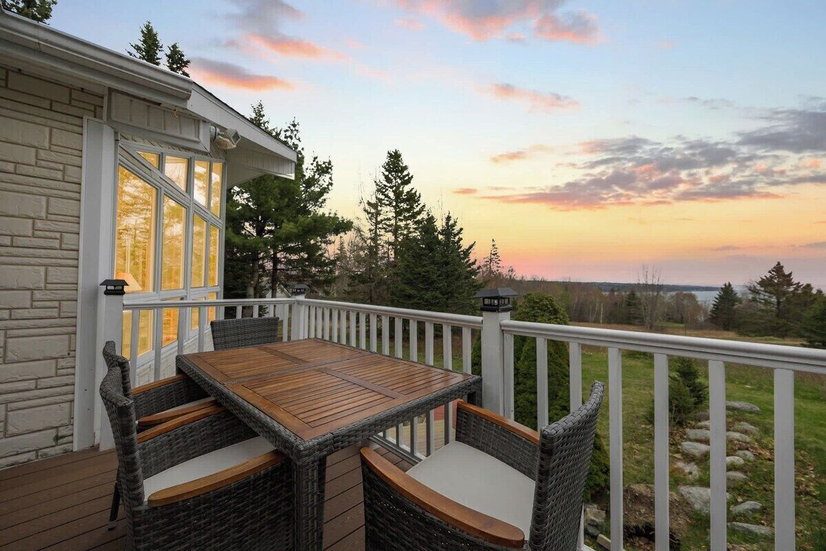 A deck with a table and chairs, overlooking a natural landscape with trees and a sunset.