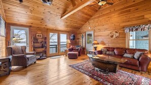 A wooden cabin interior with a sofa, armchair, and ottoman, a fireplace, and a ceiling fan. - Moose Lodge on the Lake (Ellsworth)