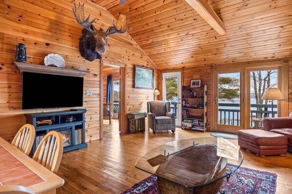 L’intérieur d’un chalet en bois est aménagé avec un téléviseur, des étagères à livres, une cheminée et une grande fenêtre donnant sur un plan d’eau.