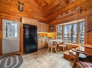 A wooden cabin interior with a kitchen, dining area, and a window overlooking a snowy landscape.
