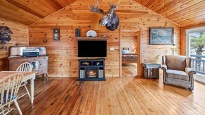 A log cabin interior with a mounted deer head, a fireplace, a television, and a dining area. - Moose Lodge on the Lake (Ellsworth)