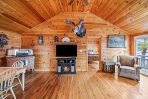 A log cabin interior with a mounted deer head, a fireplace, a television, and a dining area.