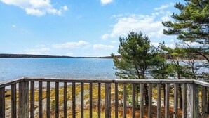 A wooden deck overlooking a calm lake with trees and a distant shoreline. - Berry Cove Cottage-Oceanfront! (Lamoine)
