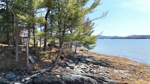 A lakeside property with a wooden deck, pine trees, and a rocky shoreline. - Berry Cove Cottage-Oceanfront! (Lamoine)
