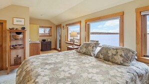 A bedroom with a large bed, a wooden cabinet with bowls, a lamp, and a view of the water. - Berry Cove Cottage-Oceanfront! (Lamoine)