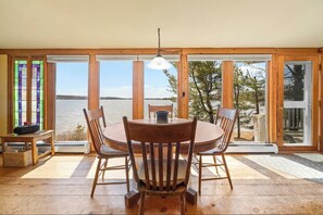 A wooden dining table with chairs, a vase, and a lamp in a room with large windows overlooking a body of water.