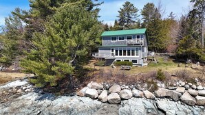 A two-story cabin with a green roof, surrounded by a rocky landscape and dense trees. - Berry Cove Cottage-Oceanfront! (Lamoine)