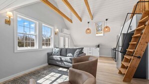 A spacious loft with a high ceiling, exposed wooden beams, a gray sofa, a brown armchair, a kitchen area, and a wooden staircase. - Cove & Cottage at Newbury Neck (Surry)