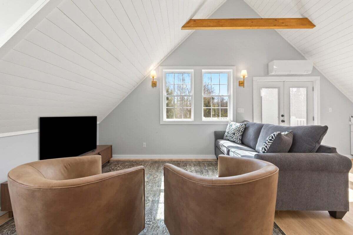 Une pièce de style loft avec un plafond en pente, un canapé gris, deux fauteuils marron, un téléviseur à écran plat et une fenêtre agrémentée de deux appliques murales.