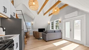 A modern kitchen with a stove, refrigerator, and wooden beams. There is a gray sofa and a dining table with chairs. - Cove & Cottage at Newbury Neck (Surry)