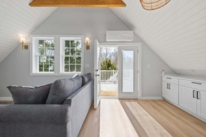 Ein Zimmer mit grauem Sofa, weißen Schränken, einer Holzbalkendecke und Blick durch eine Tür auf einen Außenbereich.
