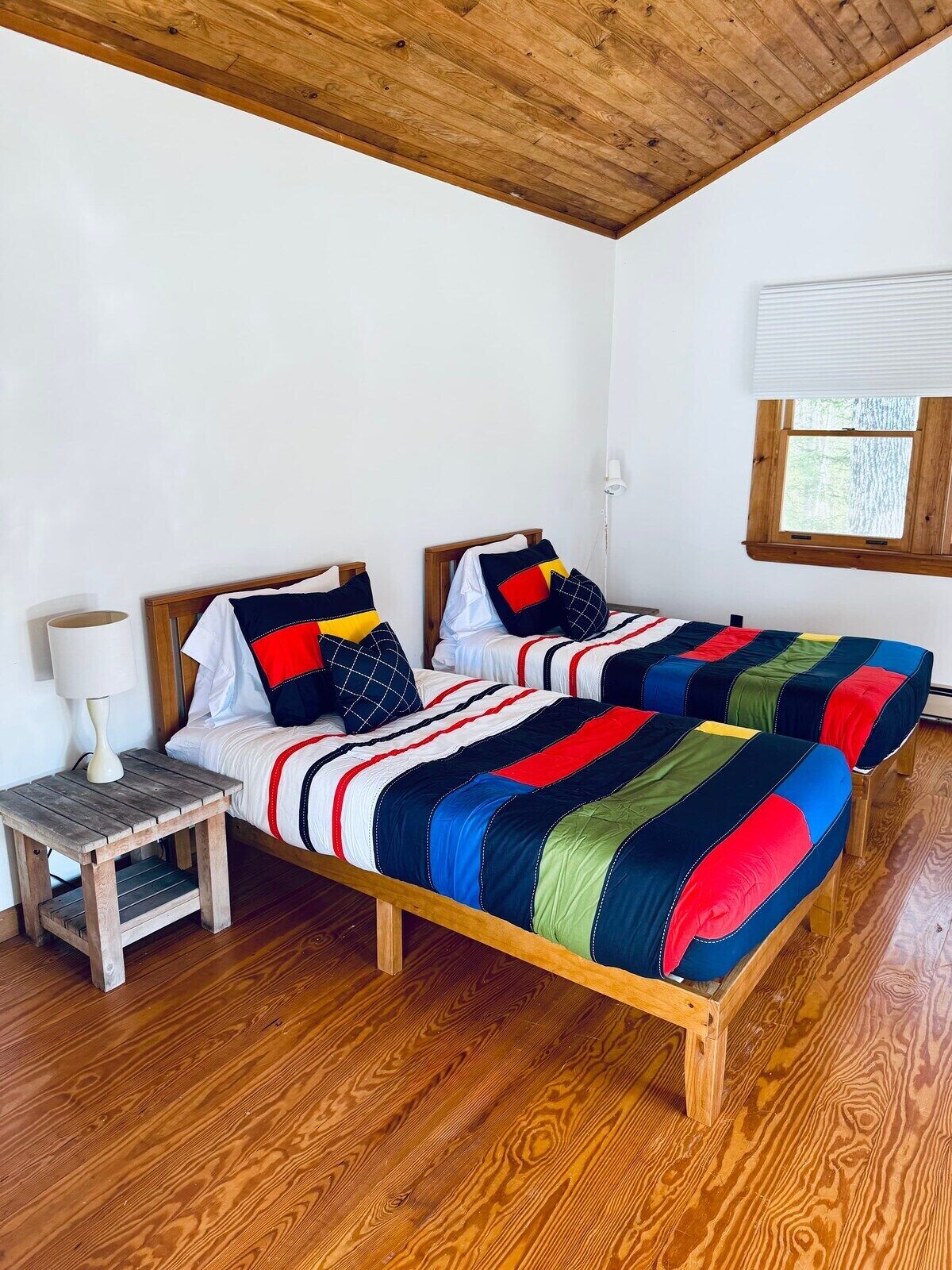 Une chambre à coucher avec deux lits en bois, une table de chevet, une lampe et une fenêtre avec des stores.