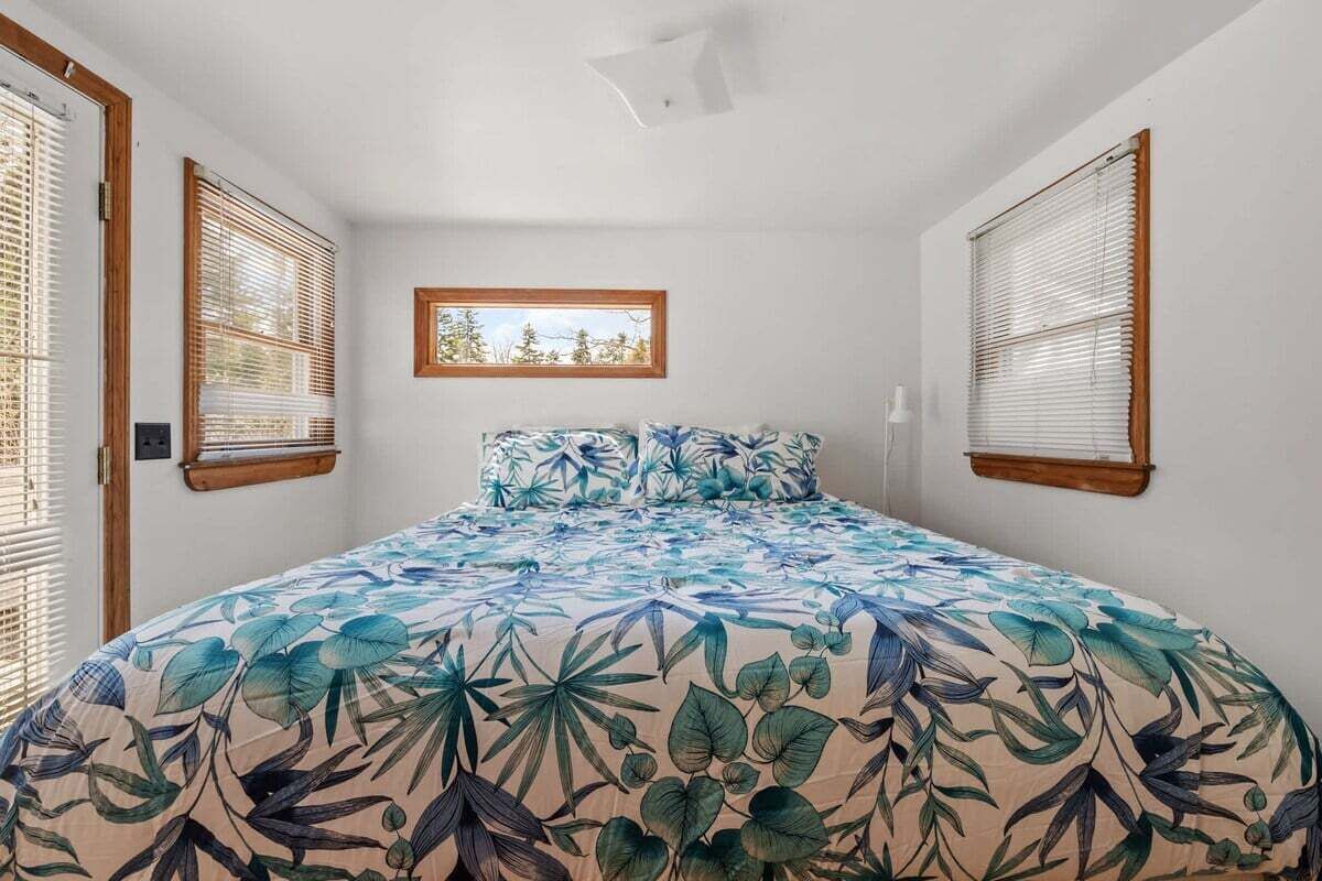 A bedroom with a bed, two windows with blinds, a small picture frame on the wall, and a ceiling light fixture.