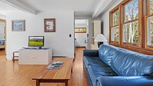 Un soggiorno con un tavolino in legno, un divano in pelle blu, una televisione su un mobile bianco e ampie finestre con vista sugli alberi.