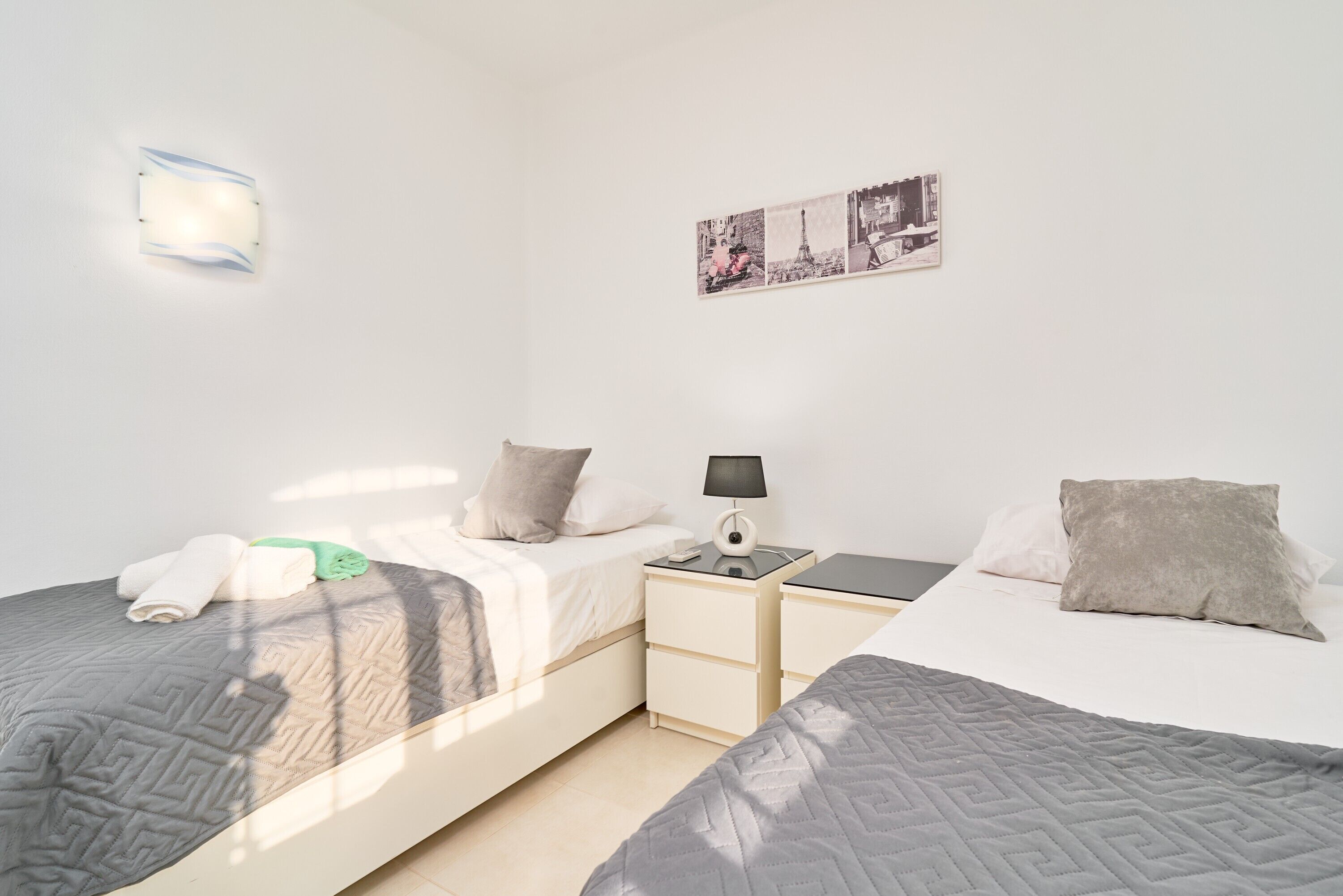 Ein Hotelzimmer mit zwei Betten, einem Nachttisch mit Lampe, einer Wandleuchte und einem Bild von Paris.