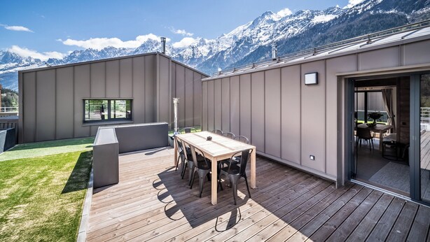 Un patio exterior moderno con una mesa y sillas de madera, una zona de barbacoa y vistas a montañas nevadas.