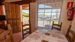 A dining area with a table set for two, wooden chairs, a window with a view of parked cars and a hill, and a fire extinguisher on the wall. - Les Xanes: El Trasgu - mountain views, terrace, and Wi-Fi (Cangas de Onís)