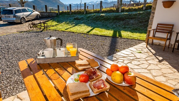 A breakfast setup with bread, ham, tomatoes, and fruit on a wooden table, with a scenic mountain view in the background. - Les Xanes: El Trasgu - mountain views, terrace, and Wi-Fi (Cangas de Onís)