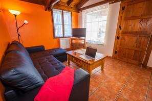 Una sala de estar con sofá, una mesa de centro con una computadora portátil, televisor y ventana con cortinas.