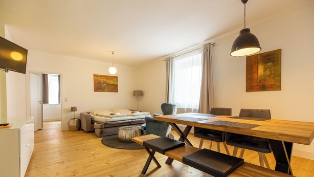 Ein modernes Wohnzimmer mit einem Holzesstisch, Bänken und einer Couch. Ein Fernseher ist an der Wand montiert und über der Couch hängt ein Gemälde.
