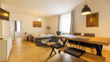 Ein modernes Wohnzimmer mit einem Holzesstisch, Bänken und einer Couch. Ein Fernseher ist an der Wand montiert und über der Couch hängt ein Gemälde.