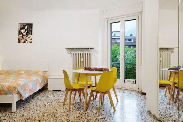 A room with a bed, a table with chairs, a radiator, and a window with a view of greenery.