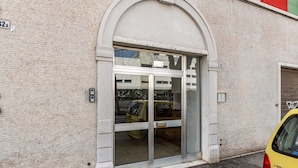 Ein Gebäudeeingang mit Glastür, ein gelber Wagen parkt davor, und eine Ziegelwand ist zu sehen.