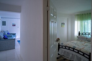 A bedroom with a bed, a window with curtains, a bedside table, and a chair in the adjacent room. - The Carhampton Villa (Montego Bay)